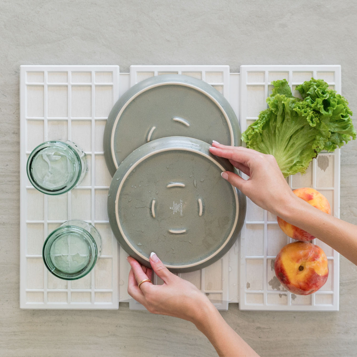 Two hands holding ceramic plates on a white modern dish pad with fruits and glasses in the background.