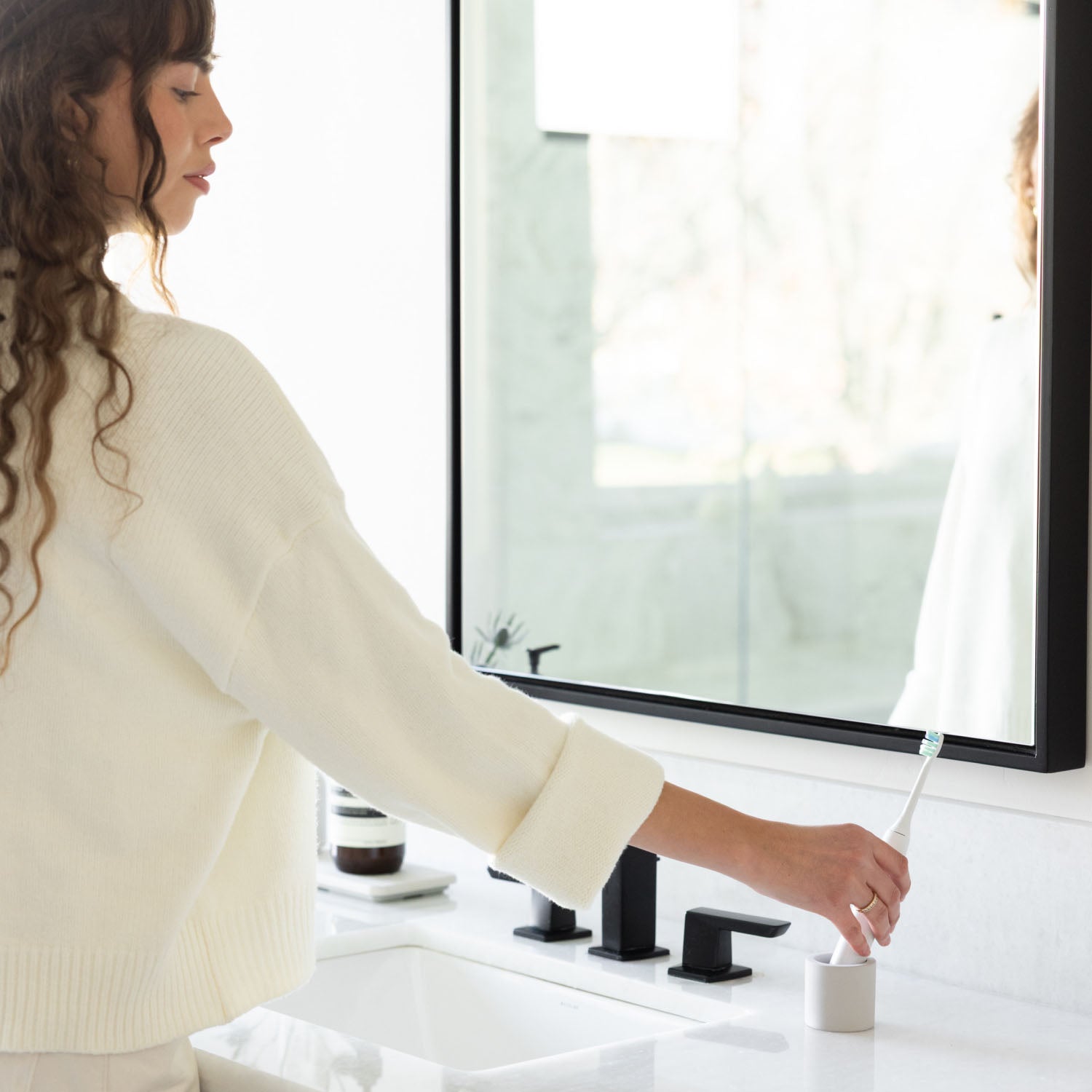 Woman placing an electric toothbrush into a stone toothbrush holder with a bathroom setting in the background.