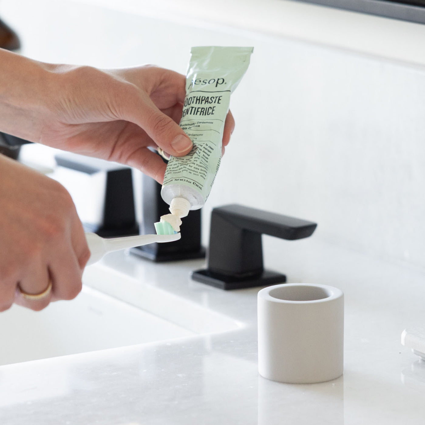 Person applying toothpaste onto a toothbrush next to a stone toothbrush holder next to a bathroom sink