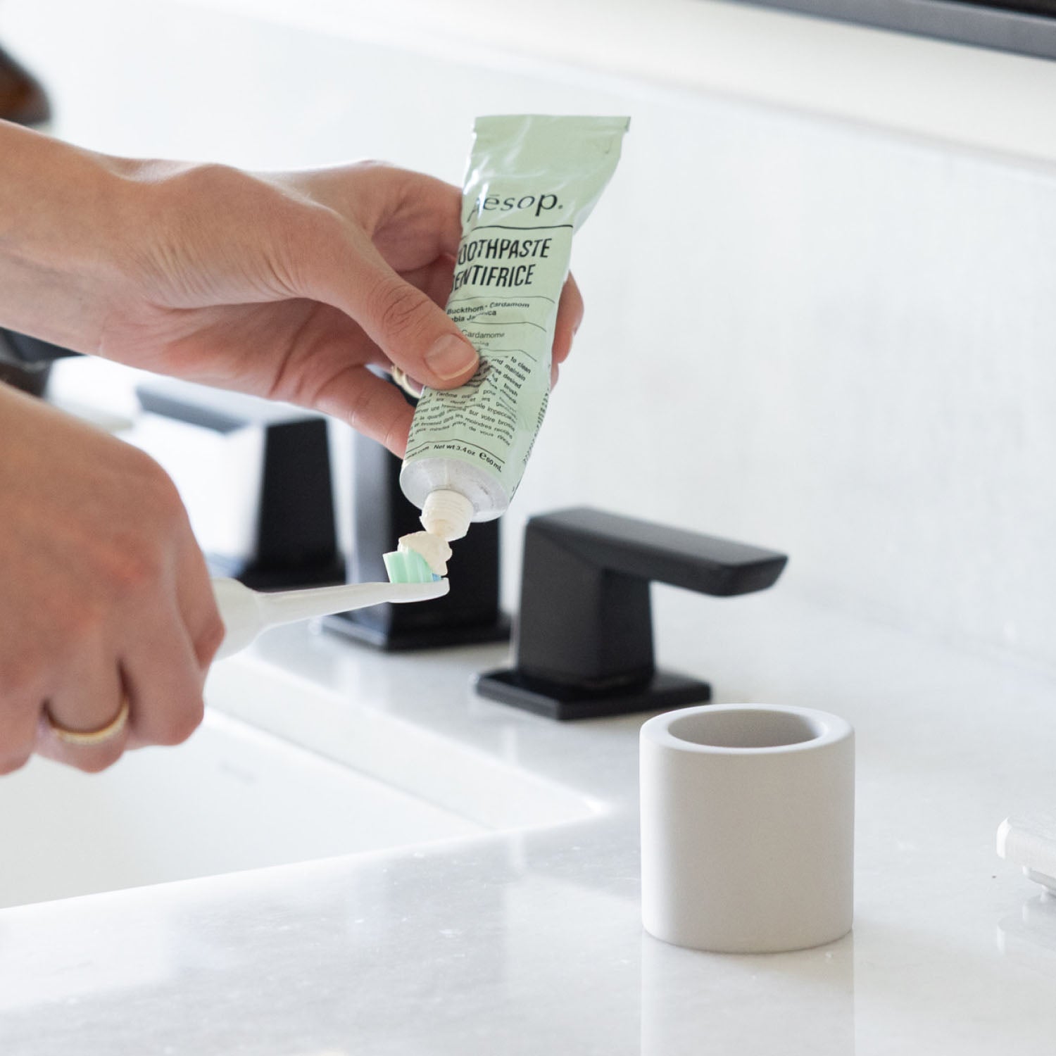 Person applying toothpaste onto a toothbrush next to a stone toothbrush holder next to a bathroom sink