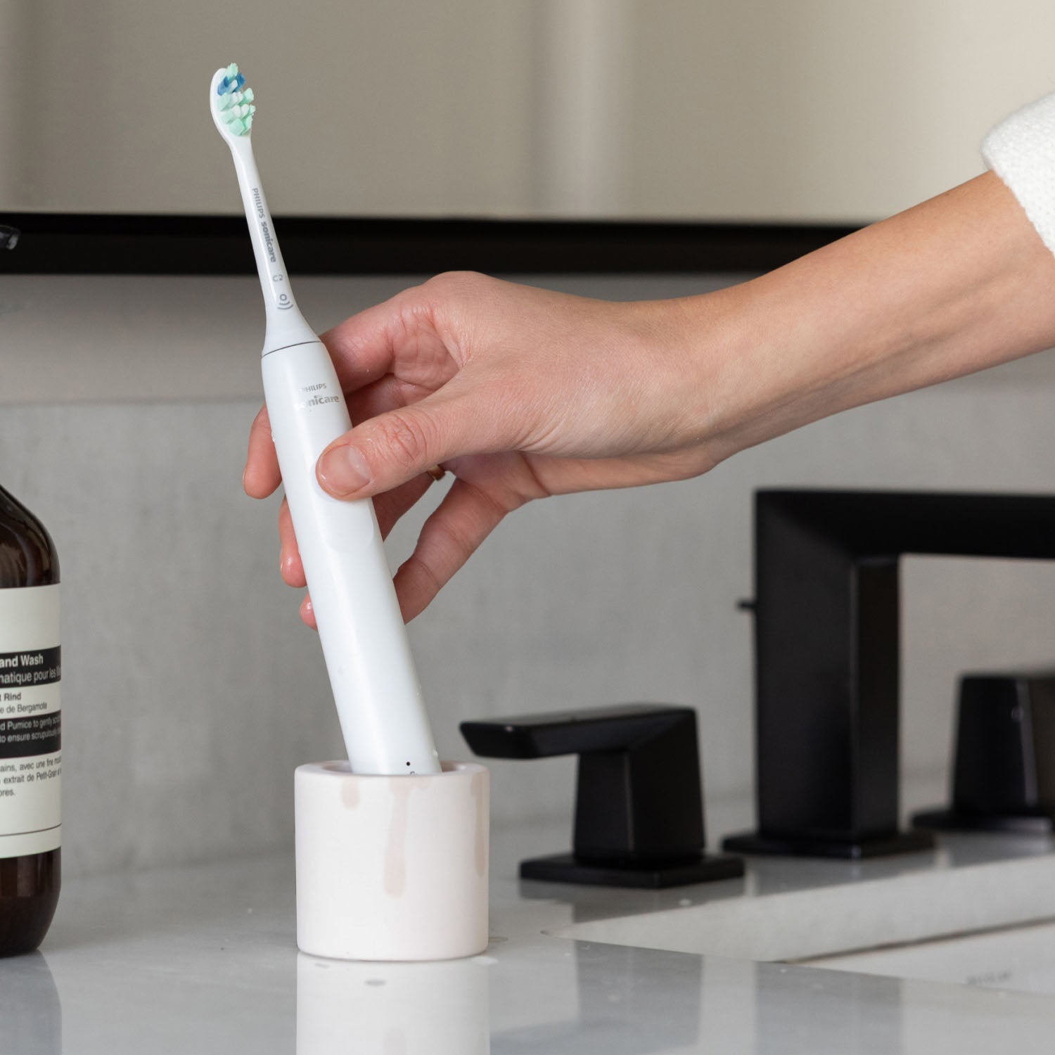 Hand placing an electric toothbrush into a stone toothbrush holder with a bathroom setting in the background.