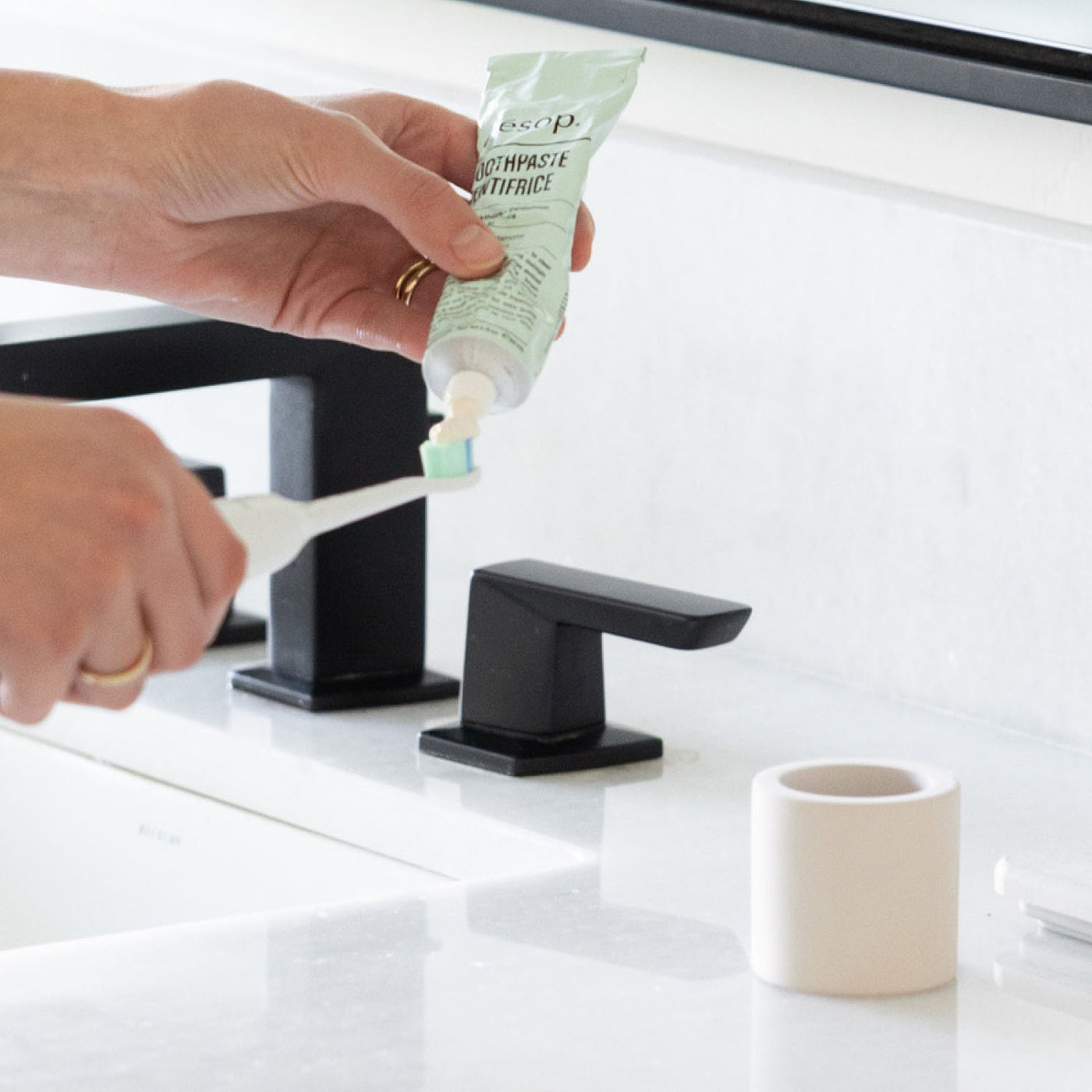 Person applying toothpaste onto a toothbrush next to a stone toothbrush holder next to a bathroom sink
