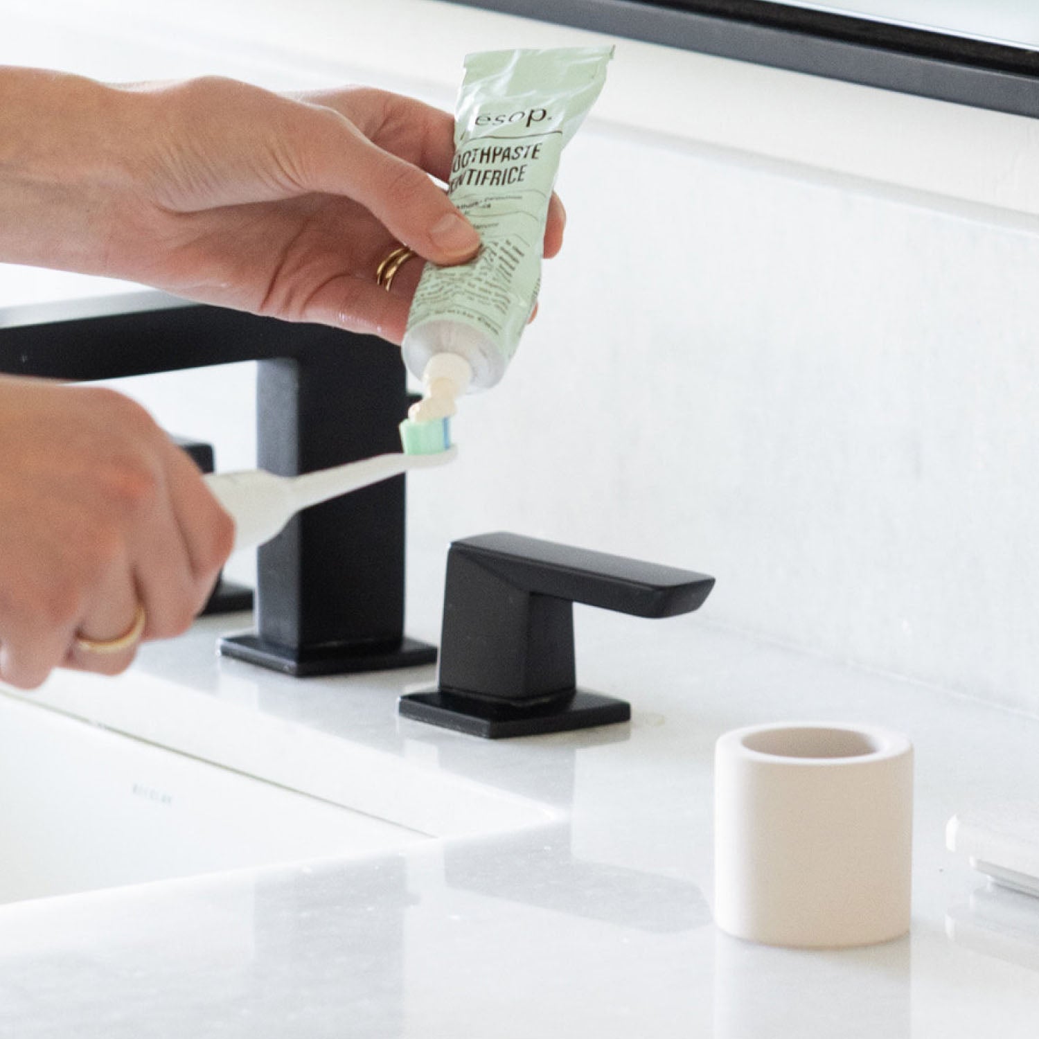 Person applying toothpaste onto a toothbrush next to a stone toothbrush holder next to a bathroom sink