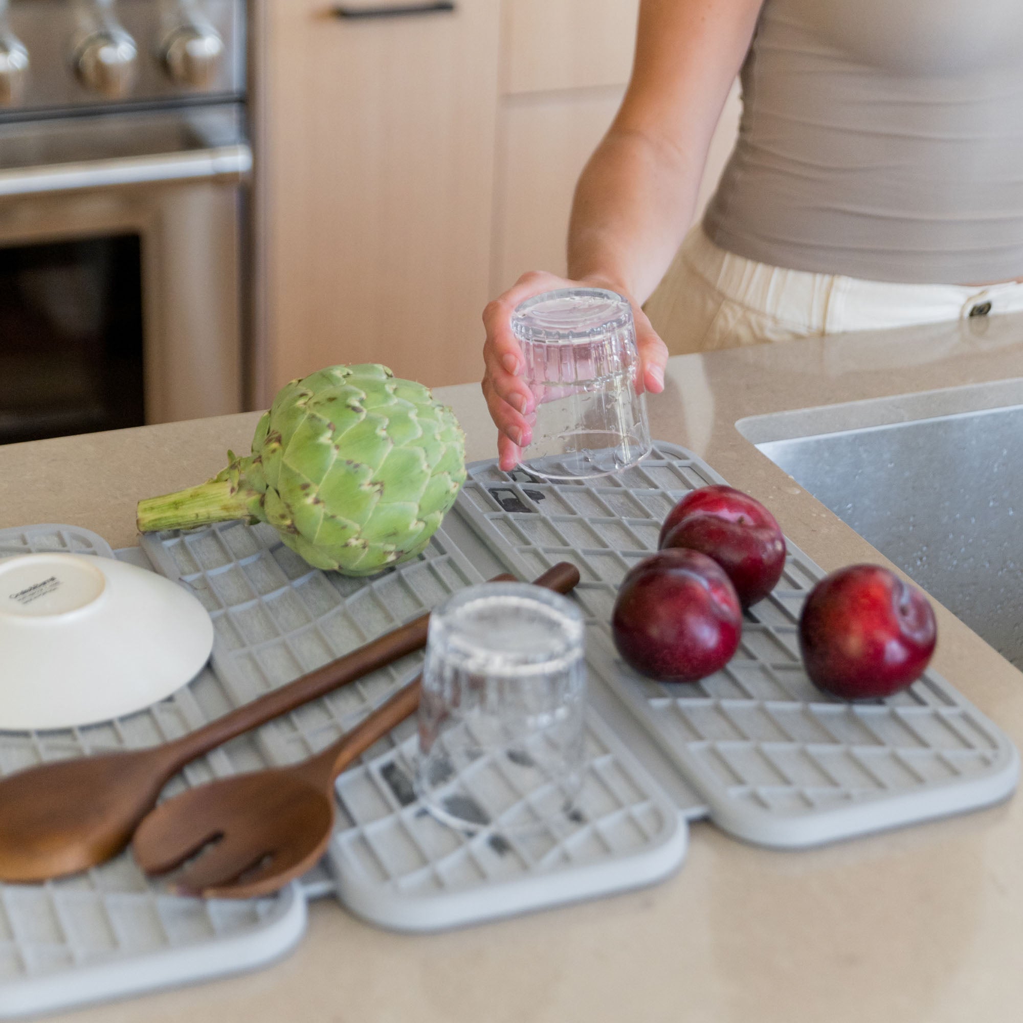Woman with Drift Ash Dish Pad with produce, pots, and glasses on it in beautiful kitchen.

